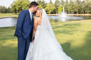Couple in wedding attire standing by lake with fountain in background.