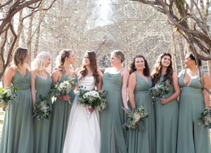 A bridal party poses outdoors wearing sage green bridesmaid dresses with bouquets of flowers.