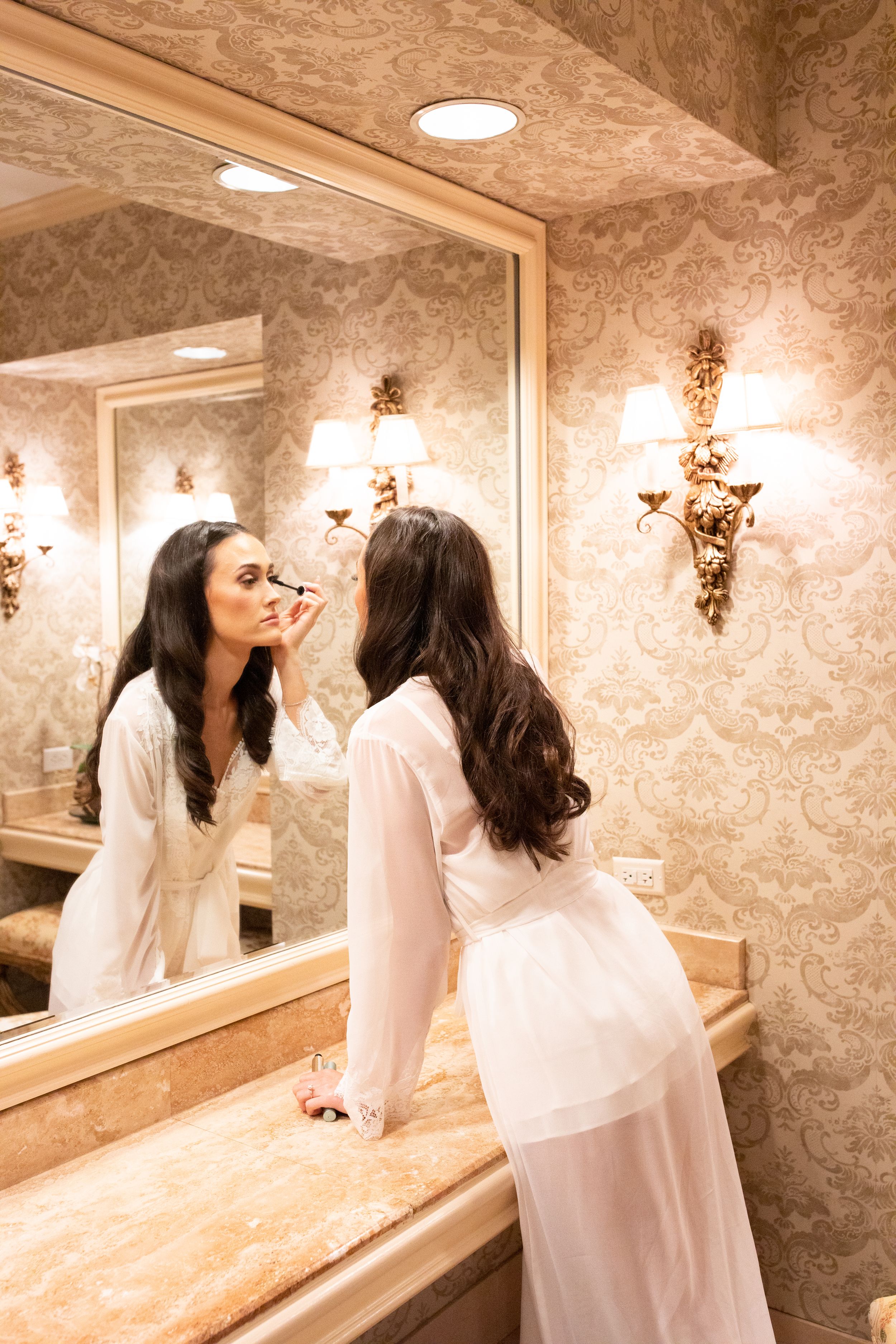 Person applying makeup in elegant bathroom with ornate golden mirrors and lighting.