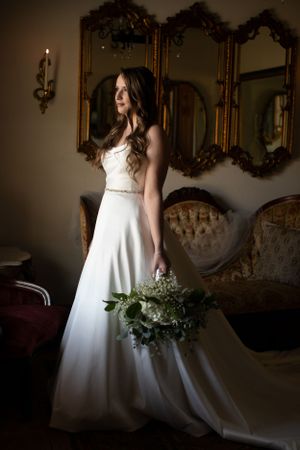 An elegant bridal portrait in a white gown standing beside ornate gold mirrors in a vintage setting.