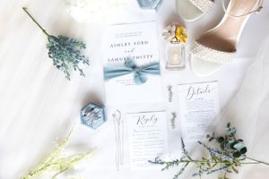Wedding stationery flatlay with blue floral details, perfume bottle and invitation cards on white fabric.