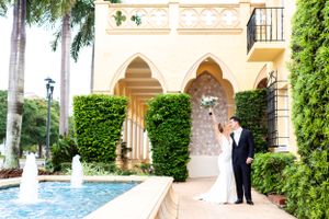 A couple shares a romantic moment by an elegant Mediterranean-style fountain and archway surrounded by palm trees.