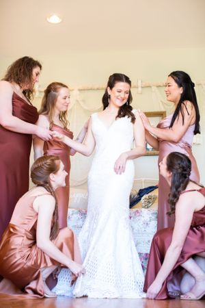 A group helps a bride get ready in her white lace wedding dress while wearing burgundy bridesmaid dresses.