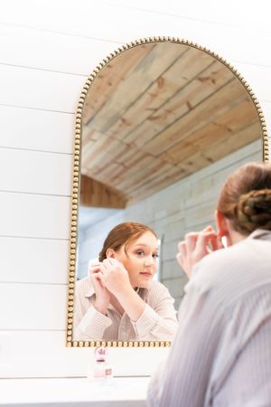 Woman putting on earrings while looking in an arched gold mirror against white shiplap walls.