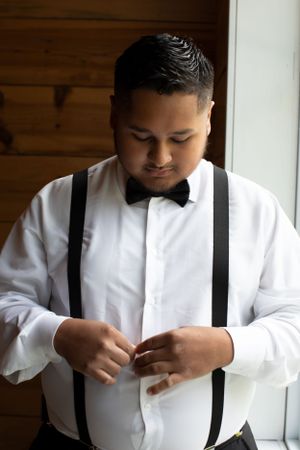 Groom adjusts bowtie while wearing white shirt with black suspenders near window.