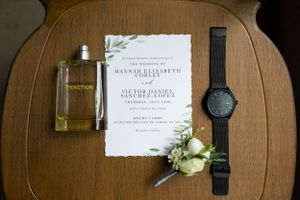 Wedding details flatlay with perfume bottle, invitation, watch and boutonniere on wooden surface.