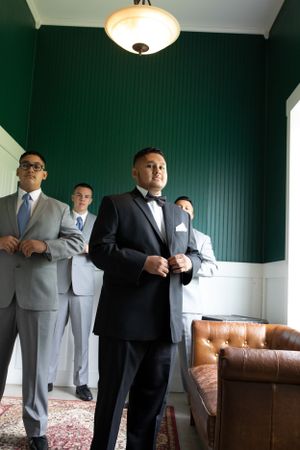 Groomsmen in mixed formal attire stand in room with dark green walls and vintage lighting.