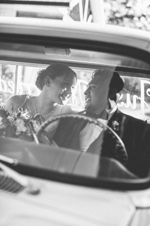 A black and white photo showing newlyweds sharing an intimate moment inside a vintage car.