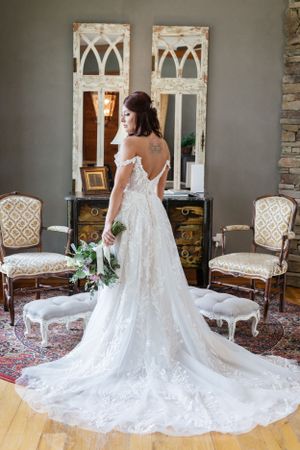 Elegant wedding dress with long lace train displayed in vintage-styled room with ornate chairs and mirrors.