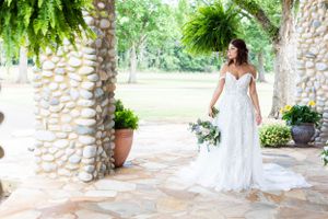Bride in white lace gown poses on stone patio with hanging ferns and garden backdrop.