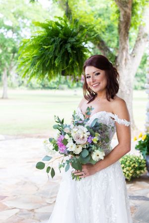 Bride in off-shoulder lace wedding dress holds vibrant purple and white bouquet under hanging ferns in garden setting.