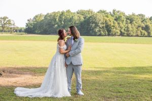 Couple in wedding attire embracing in a sunlit field with trees in background.