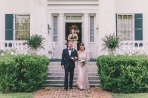 A wedding couple standing on the front steps of a white colonial-style house with green shutters.