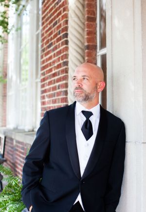 A formal portrait showing a person in a classic black tuxedo standing against a brick wall.