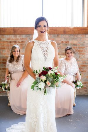 A bride in a lace wedding gown holds a bouquet while bridesmaids in pink dresses sit behind her.