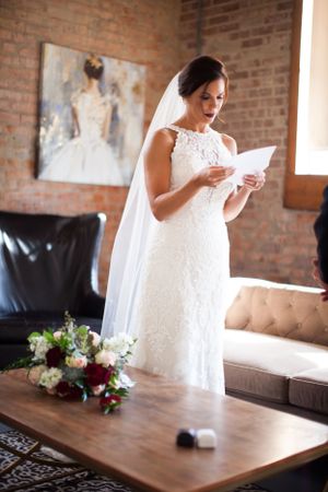 A bride in a lace wedding dress reads a letter while standing in a rustic brick room.