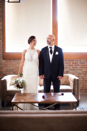 Wedding couple stands together in elegant attire in front of exposed brick wall and modern furniture.