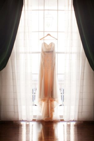A peachy-blush wedding gown hangs elegantly in front of a sunlit window with dark curtains on either side.