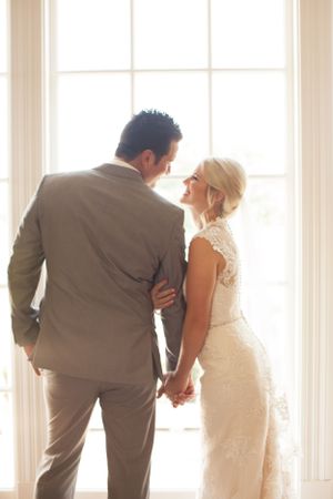 Romantic moment between newlyweds standing by bright window in formal attire.