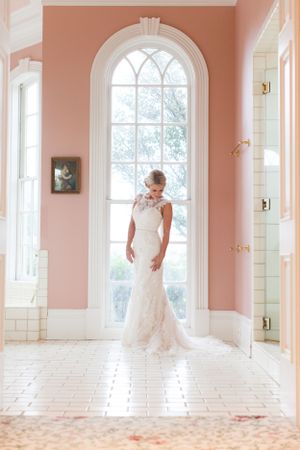 Bride in fitted lace gown standing in elegant coral-colored room with arched window.
