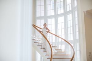 Bride in white gown posed on curved wooden staircase with tall windows.