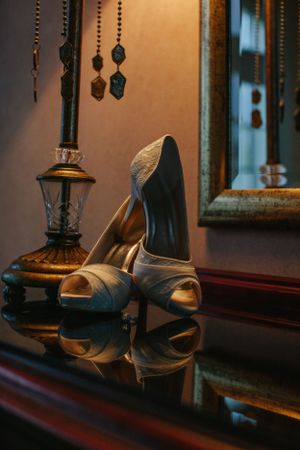 Elegant silver peep-toe bridal shoes displayed next to vintage lamp on dark wooden surface.