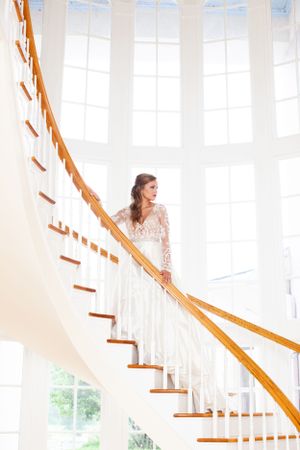 Elegant bride in lace gown poses on white spiral staircase with natural lighting.