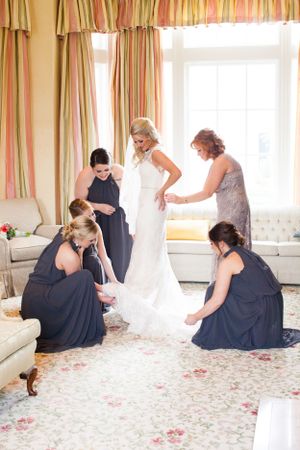 A bridal party helps adjust a white wedding dress in an elegant room with striped curtains.