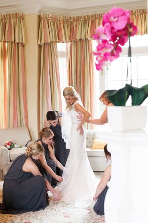 Bridal party helps adjust wedding dress in elegant room with striped curtains.