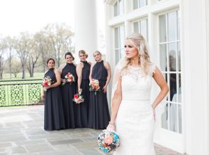 Bride in white gown with bridesmaids in navy dresses holding floral bouquets on outdoor terrace.