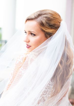 Side profile of a bride wearing a flowing veil and lace gown in natural window light.