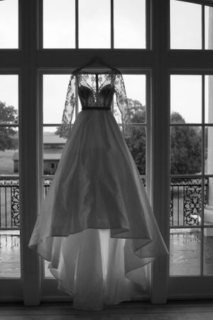 Black and white photograph of an elegant wedding gown with lace sleeves hanging in front of floor-length windows.