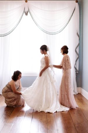 Three women help with wedding dress preparations in front of elegant white drapes.