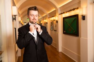 A person in formal attire adjusts their bow tie in an arched hallway.
