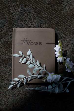 Brown leather vows book with silver decorative leaves and white flowers on dark textured background.