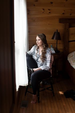 Moody portrait of someone in plaid shirt sitting by window in rustic cabin setting.