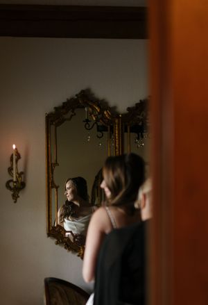 Reflection in ornate gold mirror shows preparation scene with elegant decor and candlelight.
