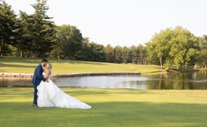 A wedding couple poses on a golf course with a serene pond and fountain in the background during golden hour.