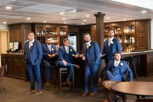 Groomsmen in matching navy suits pose casually at a wooden bar in an elegant venue.