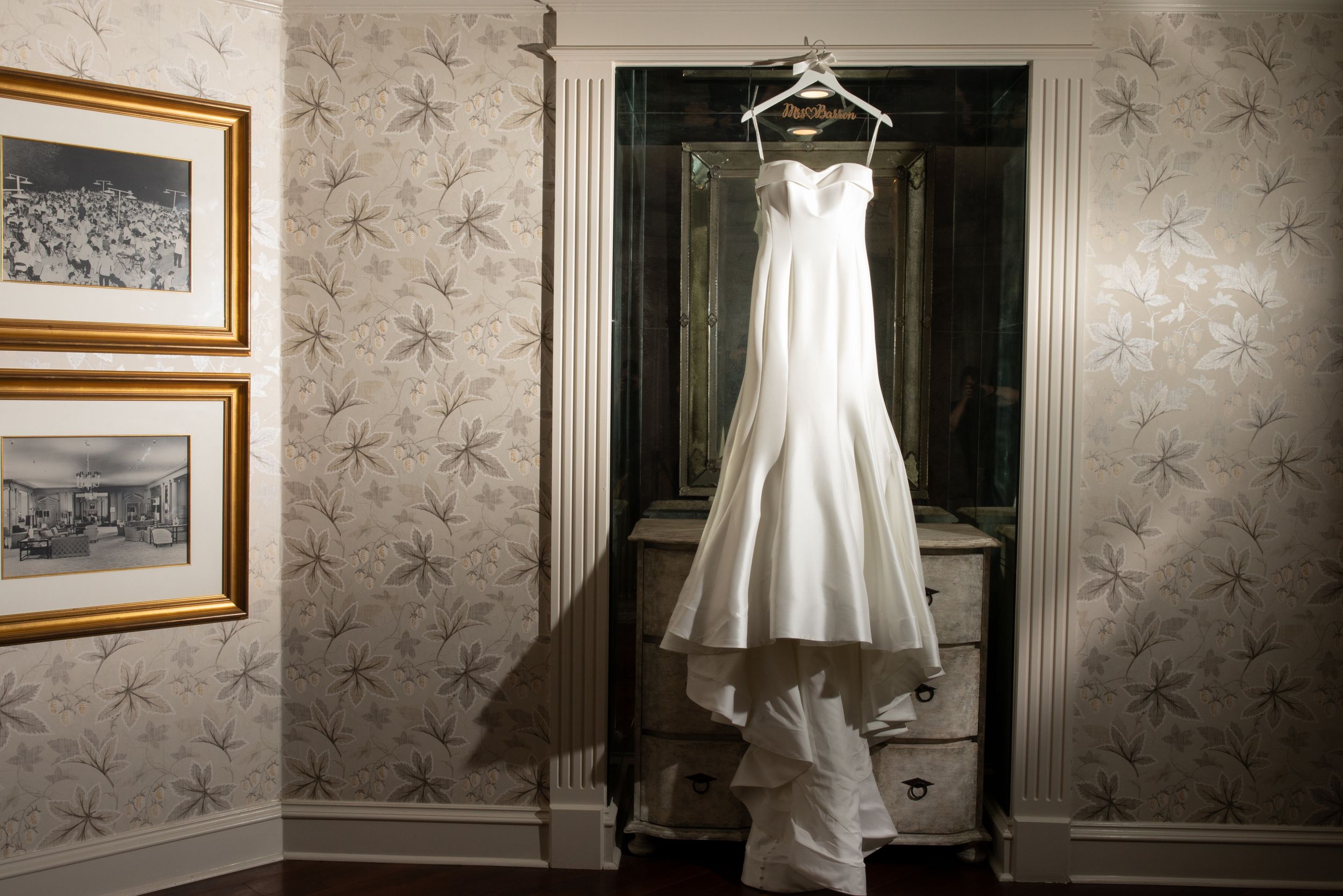 Classic white wedding dress hanging in front of floral wallpaper in elegant room.