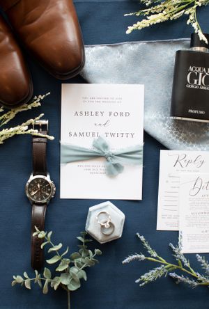 Wedding invitation with brown leather shoes, watch, and eucalyptus sprigs on navy blue background.