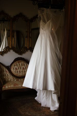 Elegant white wedding dress hanging in vintage room with ornate gold mirrors and antique furniture.