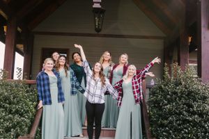 Group of bridesmaids in blue dresses and plaid shirts celebrating on rustic porch.