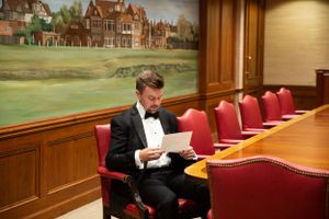 A person in a tuxedo reads documents in an elegant wood-paneled room with red chairs.