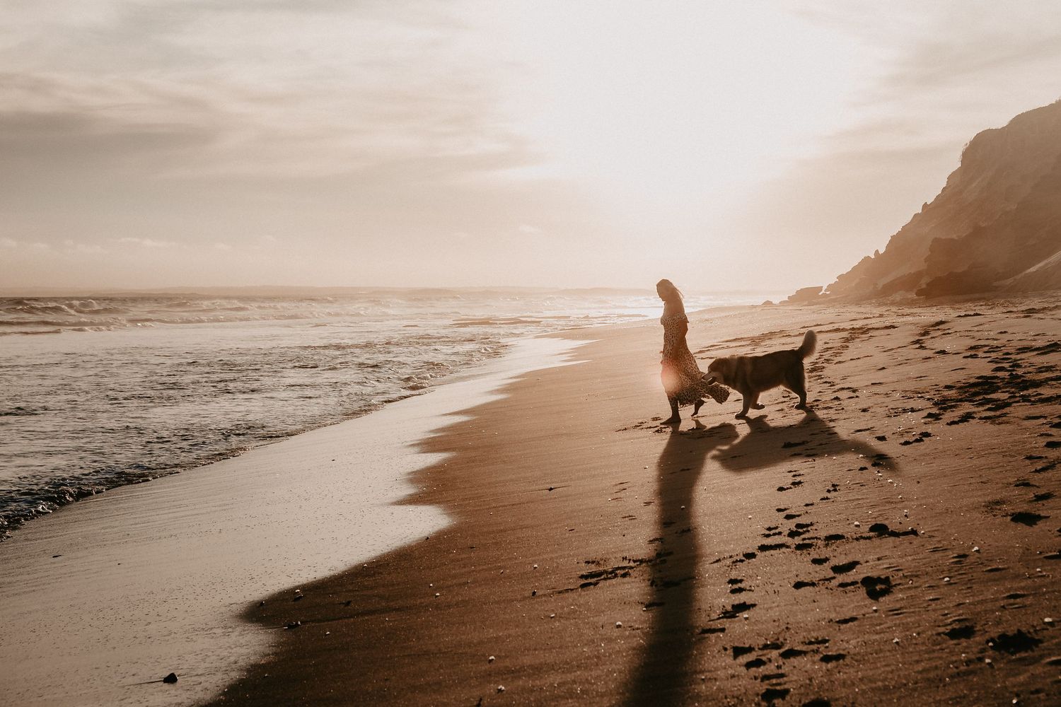 Silhouette of dog and walker against sunset on empty beach with cliffs in background.