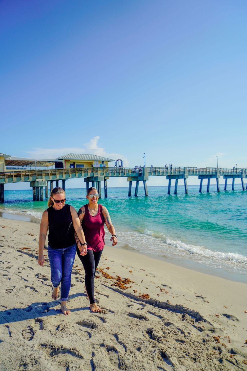 Two women walking hand in hand on a sunny beach with a pier in the background.