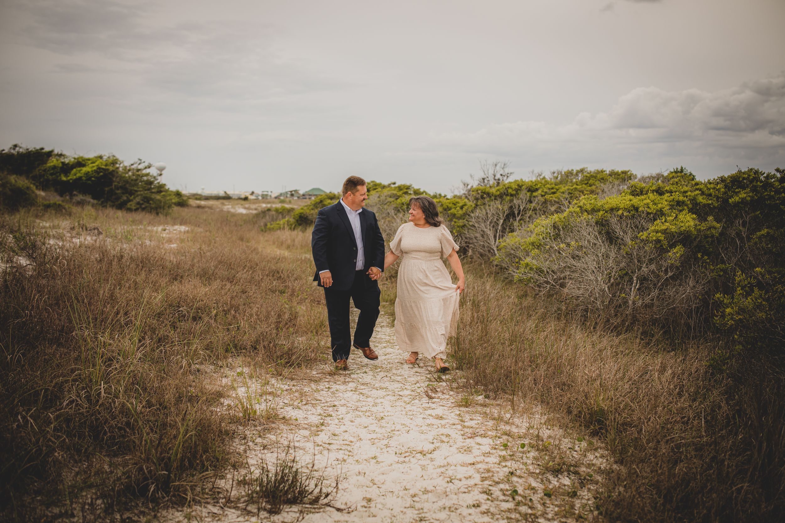 A couple walks hand in hand along a sandy coastal path surrounded by natural vegetation.