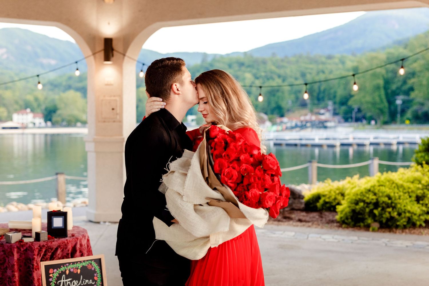 Couple shares intimate moment on lakeside terrace with red roses and mountain backdrop.