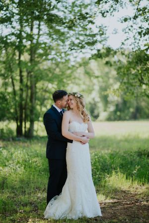 Bride in lace gown and groom in navy suit embrace in a romantic woodland setting.
