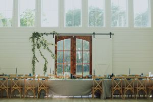 White reception venue with rustic wood doors, cross-back chairs, and elegant greenery decorations.